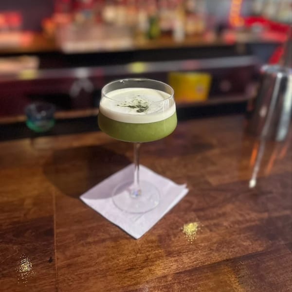 A green cocktail with white foam and dark sprinkles sits on a napkin at a bar.