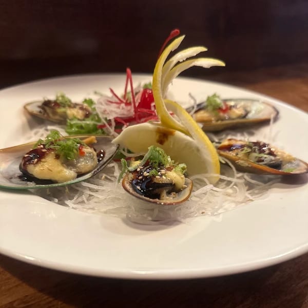 Five baked mussels with sauce, sesame seeds, and greens, garnished with lemon and radish on a white plate.