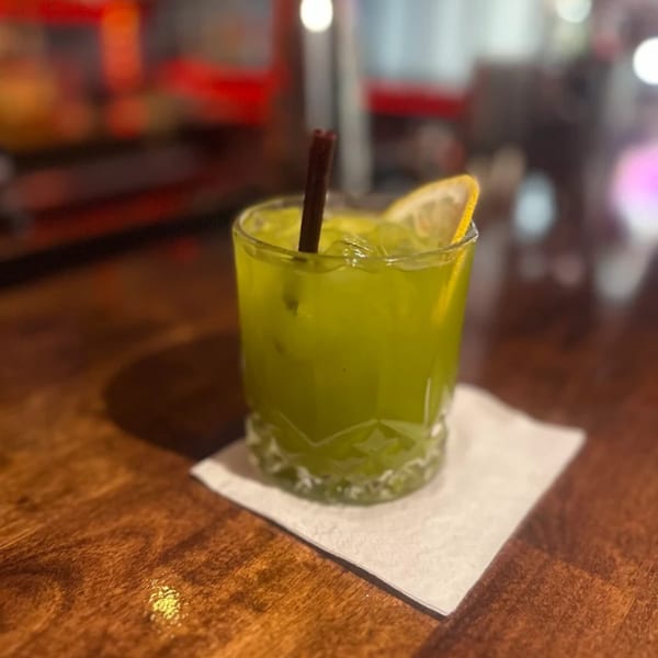 A vibrant green cocktail with ice, a lemon slice, and a cinnamon stick on a wooden bar.