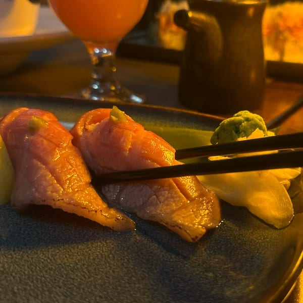 Chopsticks picking up seared salmon nigiri sushi with wasabi and pickled ginger on a dark plate.