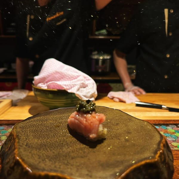 Close-up of a single piece of Yamashiro sushi with caviar on a dark plate, chef in background.