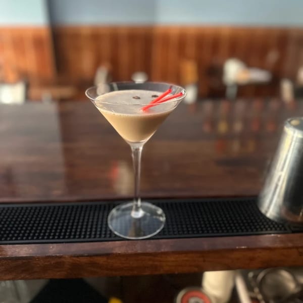 A creamy martini with coffee beans and red straws sits on a dark wooden bar.