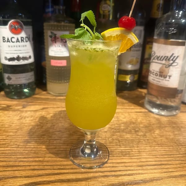 A vibrant yellow cocktail garnished with mint, an orange slice, and a cherry, on a wooden bar.